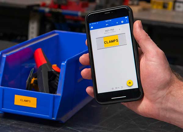 Ein gestaltetes Etikett auf dem Bildschirm der mobilen App „Express-Etiketten“ neben dem gedruckten Etikett. Beide haben eine auffällige gelbe Farbe und den Text „CLAMPS“.