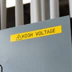 Ein großes gelbes Brady-Etikett mit der Aufschrift „High Voltage“ (Hochspannung) ist an einer elektrischen Schalttafel angebracht.